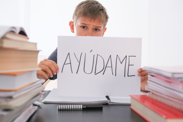 Upset tired boy sitting at the table with many books, exercises books. Spanish word Auydame - Help...