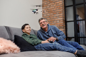 Old grandfather and little boy grandson play electronic toy plane lay on sofa, funny small grandkid...