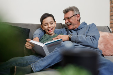 Smiling grandson reading book with his happy gray grandfather while sit on sofa