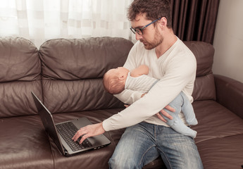 Fototapeta premium Father working at home using a laptop with a little baby on his hands. Freelancer. Stay at home concept.