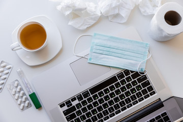 A laptop with a white cup of tea and saucer on quarantine, a medical mask on the keyboard, a roll of toilet paper and used napkins, tablets and a thermometer. All this is located on a white table