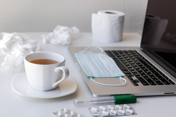 The laptop with a white cup of tea and saucer on quarantine, a medical mask on the keyboard, a roll of toilet paper and used napkins, tablets and a thermometer. All this is located on a white table.