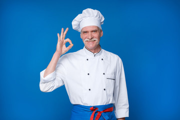 Portrait of elderly master chef isolated on blue background, with okay gesture
