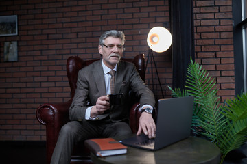 A millionaire in a business suit with a cup of coffee in his hands solves business issues on the laptop of his stylish office. Business lifestyle.