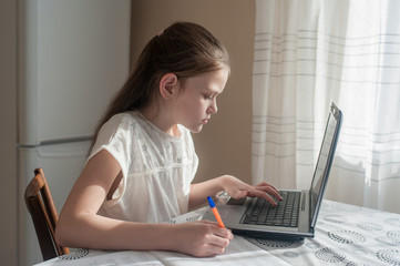 A school-age girl sits at home at a table and studies using a laptop. Distance Education, Quarantine