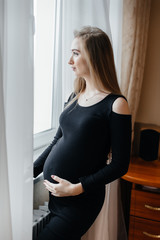 A beautiful pregnant girl is standing in a room near the window