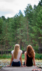 Two women are sitting together on pier enjoying the scenic view of a lake,back view. Female best friends enjoying a day.Beautiful romantic sunset image/Two girls admires the beautiful landscape