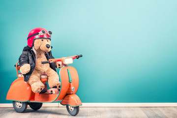 Retro Teddy Bear toy in red helmet with goggles and leather jacket on old children's pedal orange scooter from 60s front mint blue wall background. Kid's race concept. Vintage style filtered photo