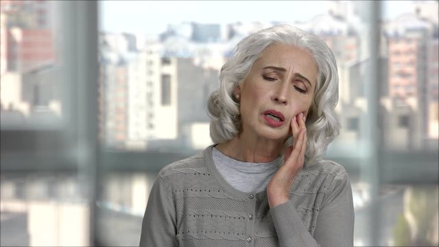 Old woman having toothache. Senior lady with pain caused by gum disease, touching cheeck and suffering. Indoor, bright office with cityscape view.