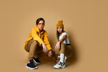 Young teens boy and girl in comfortable clothing, hats, sneakers and sunglasses sitting and pointing at camera