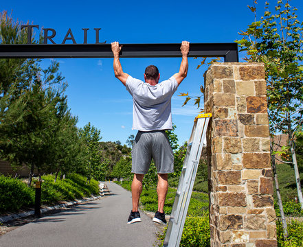 Man using a steel beam of a park sign in order to perform pull ups as part of a work out.  A ladder is visible nearby