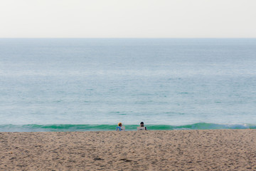 Two people sitting on a sandy beach in front of green wave coming out of blue ocean