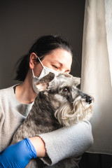 Sick woman with mask and medical gloves hugs a dog while looking through the window