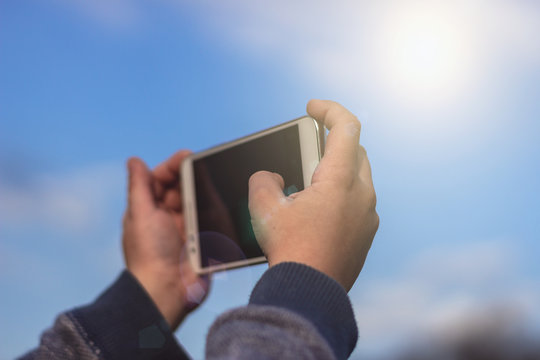 Little boy or a girl holding a smartphone using both hands and taking a photo of the blue sky. Colorful sunny outdoors background, travel concept, close up.