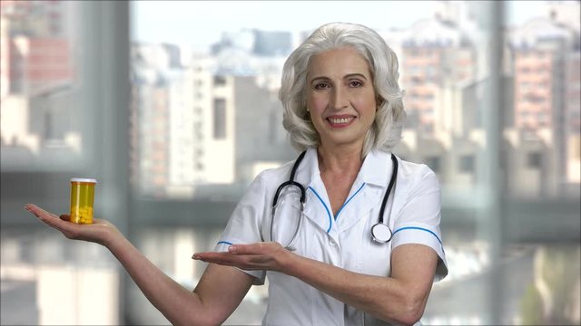 Senior doctor lady shows bottle of pills. Adverticing and promotion, windows with city view background.