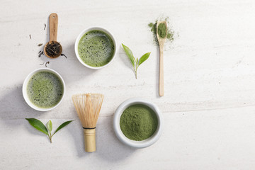Organic matcha green tea powder in bowl with wire whisk and green tea leaf on white stone table,...