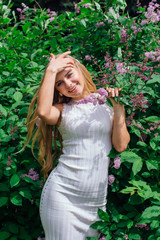 Portrait of a charming blond woman wearing beautiful white dress standing next to lilac bush.