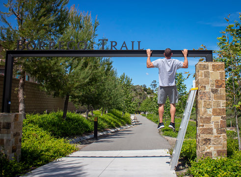 Man using a steel beam of a park sign in order to perform pull ups as part of a work out.  A ladder is visible nearby