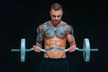 Tattooed strong muscular athletic man pumping up muscles with barbell on black background