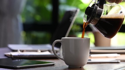 A hand pouring steaming coffee in to a cup on a work desk when work from home - Powered by Adobe