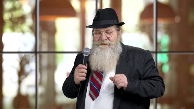 Emotional charismatic old man speaking with microphone. Senior bearded presenter in suit, warm blurred background.