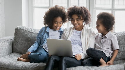 Young African mother with little daughter and son sitting on couch with laptop using on-line websites, buying choosing goods, mommy teach children online, education or having fun in internet concept