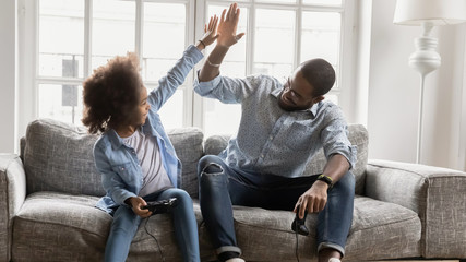 African young father and daughter hold joysticks play playstation enjoy video games sit on couch,...