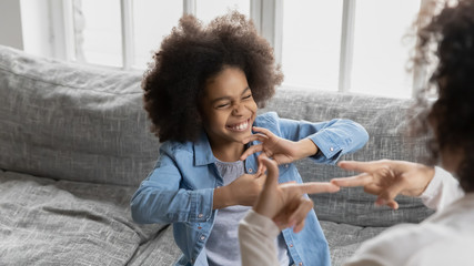 Fototapeta na wymiar African deaf kid girl and her mother sitting on couch showing symbols with hands using visual-manual gestures enjoy communication at home. Hearing loss disability sign language learning school concept