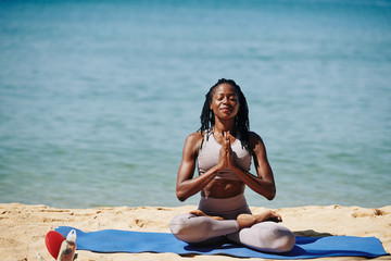 Fototapeta na wymiar Smiling Black young woman in sports top and leggings enjoying meditaton on sandy beach