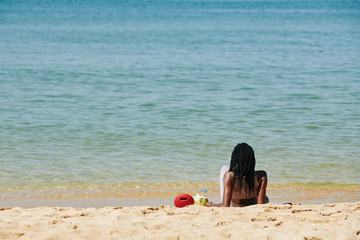 Young woman spending time on sandy beach, listening to music from portable speaker, drinking coconut cocktail and looking at waves