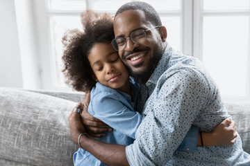 Loving African father in glasses hugs little daughter, family sitting on couch cuddling closed eyes enjoy sincere moment of tenderness, feeling bond express appreciation and gratitude for love concept