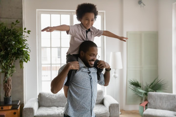 Little african preschool boy imagines himself like a airplane stretched arms seated on dad shoulders flying in air. American father spend active funny time play with kid in modern living room at home