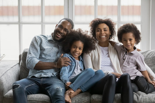 African ethnicity small siblings and parents full family portrait. Married young couple their little cute children daughter and son hugging sit on sofa in cozy light living room photo shooting at home