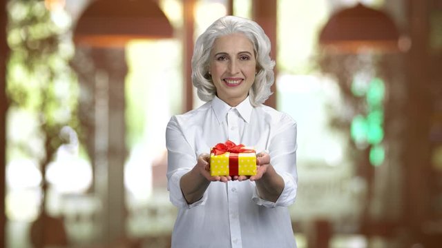 Old lady wearing white shirt showing gift box in both hands. Happy grey-haired woman, warm blurred background indoors.