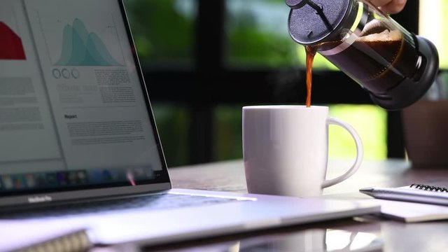 A hand pouring steaming coffee in to a cup on a work desk when work from home
