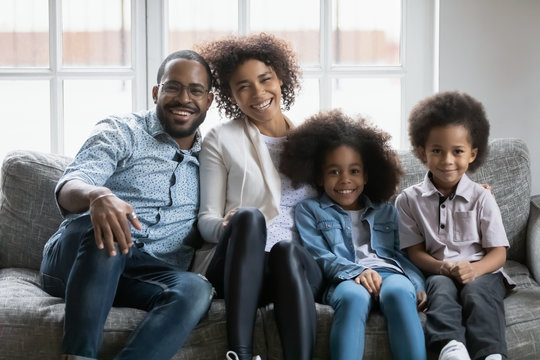 Beautiful African ethnicity full family with little son and daughter in casual wear sit on couch in living room smiling look at camera feels happy. Concept of first home, homeowners loan and mortgage