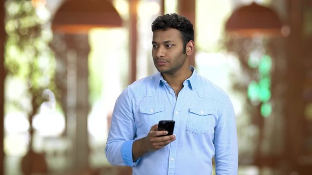 Brown-skinned office worker taking photos of someone unnoticeably. Sneaky behavior, young guy wearing blue shirt and making photos, warm blurred background.