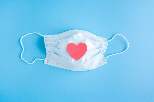 Disposable medical mask on blue background with red heart on it against bacteria and viruses. Stop the spread of coronavirus, self-defense. Copy space