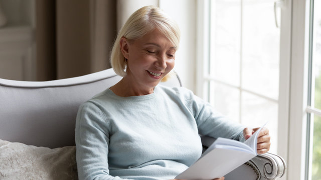 Smiling mature woman sit on couch in living room reading interesting book, happy middle-aged female relax on sofa at home enjoy novel or story, rest on leisure weekend, elderly hobby activity concept