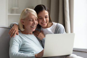 Smiling elderly mother and adult daughter sit relax on sofa in living room watch video on laptop together, happy senior mom and grown-up millennial girl child rest at home using modern computer