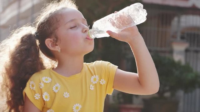 Cute girl child drinks from a plastic bottle on the street near the house. Slow motion little girl eagerly drinks drinking water. Close-up.