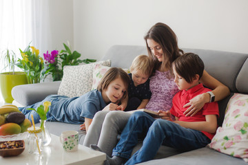 Young woman, mother with three kids, reading a book at home, hugging and laughing