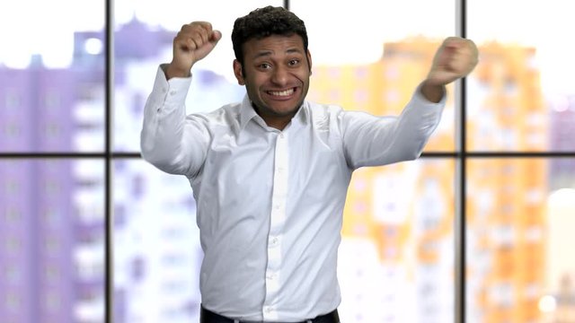 Extremely happy young dark-skinned business man wearing formal shirt. Successful worker rejoices. Window background with urban view.