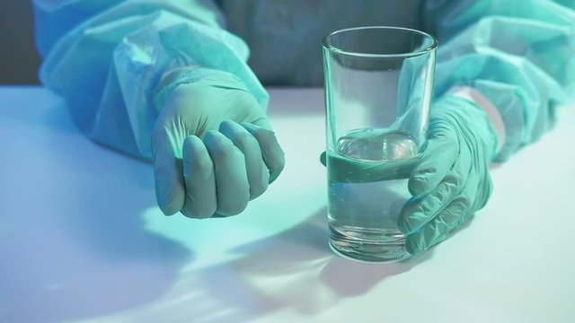 COVID-19 pandemic. Medication dosage. Doctor hands in protective gloves offering pills water glass.