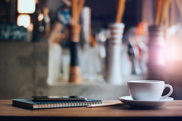 Closeup white cup of coffee with smartphone, notebook and pen on table in cafe. Vintage light, blurred and bokeh background