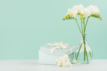 Romantic bright holiday background with spring flowers freesia in glass vase and gift box with ribbon in green mint menthe interior on white wood board.