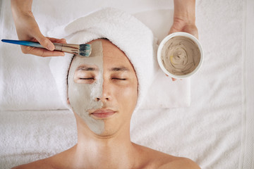 Cosmetologist applying charcoal mask on half of face of mature man, view from above