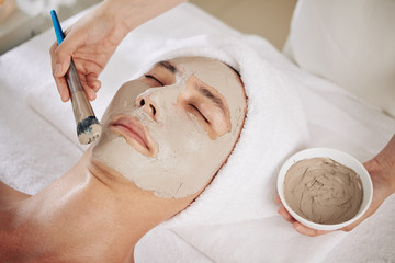 Close-up image of cosmetologist applying purifying clay mask with synthetic brush