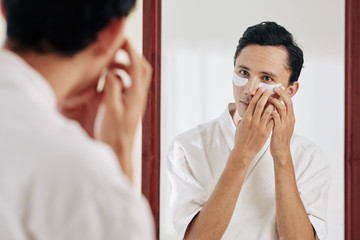 Mixed-race young man standing in front of mirror and applying undereye patches after morning shower
