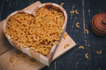 Wooden heart shaped box with pasta or raw macaroni and salt pot on black wooden background. Close up shot, copy space. Carbohydrates diet, healthy eating habits and vegetarian food concept.